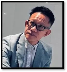 A man wearing glasses and a light blue suit jacket over a patterned shirt sits against a light-coloured wall, looking towards the camera.