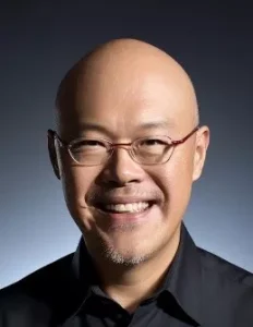 A person with short dark hair wearing a dark blazer and light shirt is smiling and posing with their right arm crossed over their chest against a plain light background.
