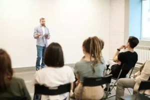 A person stands at the front of a room holding a tablet, speaking to a seated audience.