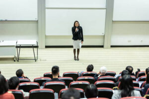 Woman speaking with a microphone in front of an audience in a lecture hall.