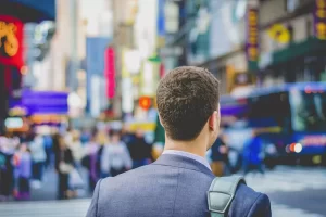 A man in a suit stands on a busy city street, facing away, with a blurred background of people, buildings, and bright signs.