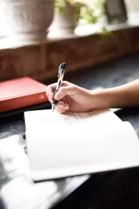 A person writing in a notebook with a pen, next to a red book and potted plants on a windowsill.