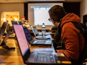 A person in a hoodie looks at a projected screen in a classroom setting, surrounded by laptops on a wooden table.