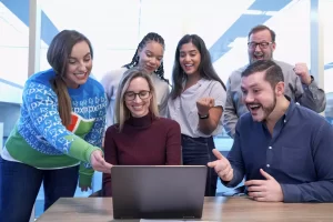 A group of six people gathered around a laptop, smiling and cheering in a bright office setting.