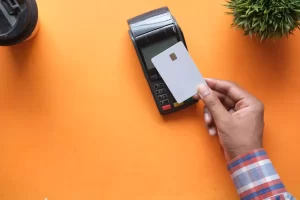 A hand holds a credit card over a payment terminal on an orange surface, with a plant on the side.
