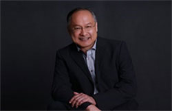 A man in a black suit is seated against a dark background, smiling at the camera.