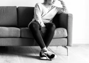 A person sitting on a gray sofa, resting their head on their hand, wearing a sweater, jeans, and sneakers in a black-and-white photograph.