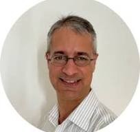 A middle-aged person with short gray hair and glasses, wearing a colorful patterned shirt, smiling at the camera against a white background.