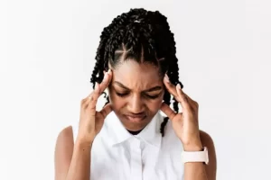 A person with braided hair and a white shirt appears stressed, pressing fingers to their temples against a plain background.