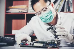 A person wearing safety goggles and a mask works on a circuit board at a desk.