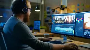 A person wearing headphones edits video footage on a computer with dual monitors in a dimly lit workspace.