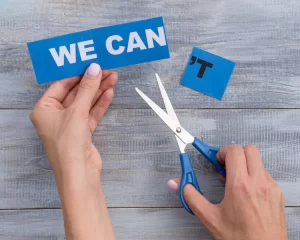 Hands holding scissors cut the word "CAN'T" into "WE CAN" on a wooden surface.
