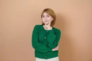 A person with short hair in a green sweater poses with one hand on their chin, against a beige background.
